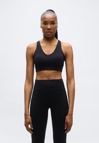 Γυμνασμένη γυναίκα με κοτσίδα, φορώντας μαύρο αθλητικό σουτιέν και κολάν με ψηλή μέση, στέκεται μπροστά σε απλό ανοιχτό γκρι φόντο.