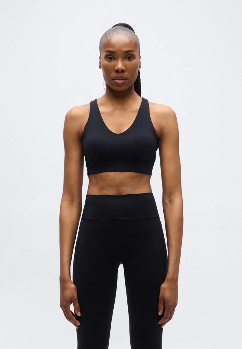 Γυμνασμένη γυναίκα με κοτσίδα, φορώντας μαύρο αθλητικό σουτιέν και κολάν με ψηλή μέση, στέκεται μπροστά σε απλό ανοιχτό γκρι φόντο.