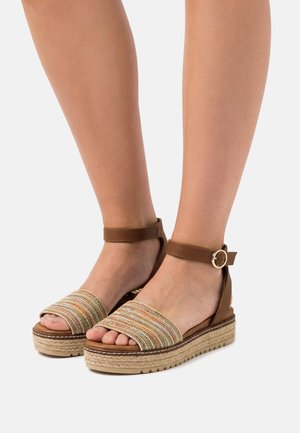 Piernas con sandalias de plataforma marrones con correas tejidas multicolores y cierres de hebilla en el tobillo sobre un fondo blanco liso.