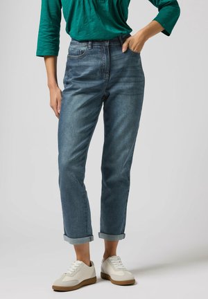 Kobieta ubrana w podwinięte niebieskie dżinsy, zieloną koszulę z długim rękawem i białe tenisówki, stojąca z jedną ręką w kieszeni na jednolitym tle.