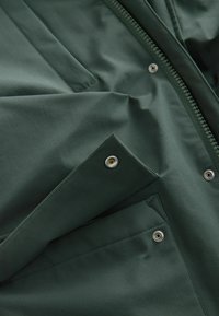 Jachetă verde cu o textură netedă, închidere prin fermoar și un buzunar cu detaliu de capac. Designul include secțiuni de țesătură suprapuse.