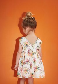 Weißes Baumwollkleid mit buntem floralem Stickmuster, V-Ausschnitt, kurzen Ärmeln und gerafftem Taillenbereich. Vor einem orangefarbenen Hintergrund.
