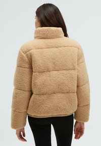 Beige fleece jacket with a high collar, boxy shape, and horizontal stitching. Textured fabric and elasticized hem. Black pants are worn.