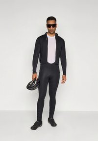Conjunto de ciclismo preto com uma jaqueta curta sobre um top claro e leggings justas. Modelo segura um capacete preto e usa óculos de sol.