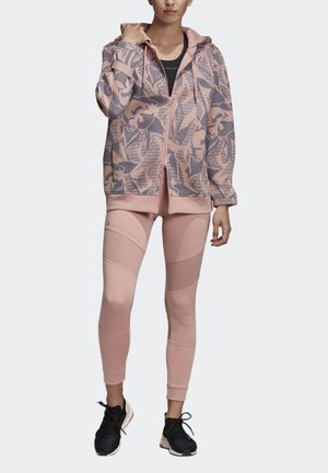 Giacca sportiva rosa con cappuccio e motivo floreale, abbinata a leggings rosa chiaro con pannelli testurizzati. Scarpe sportive nere completano l'outfit.