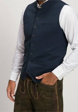Man draagt een marineblauw gilet met knopen over een wit shirt met lange mouwen en donkerbruine, geborduurde broeken in traditionele stijl, hand in de zak.