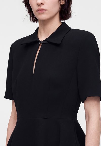 Femme portant une robe noire à manches courtes avec un petit col et une fermeture à clip sur le devant au-dessus d'une encolure en trou de serrure.