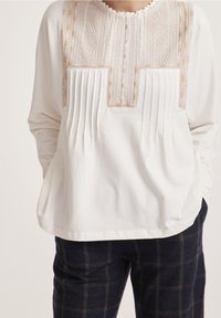 Blusa blanca de manga larga con adorno texturizado, con pliegues y cuello redondo. Se lleva con pantalones oscuros a cuadros.