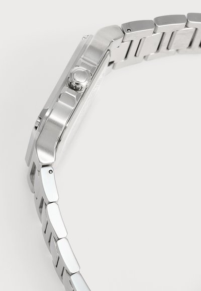 Vue latérale d'un bracelet de montre en métal argenté et d'une couronne ronde sur le boîtier de la montre sur fond blanc.