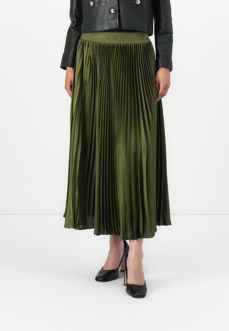 Jupe plissée verte olive avec une large ceinture-logo, en tissu brillant. Associée à des escarpins noirs et une veste en cuir noire.