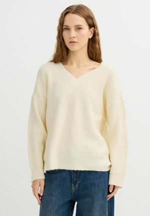 Suéter de punto color crema con cuello en V, hombros caídos, textura acanalada y dobladillo ligeramente deshilachado. Combinado con jeans de mezclilla oscuros.