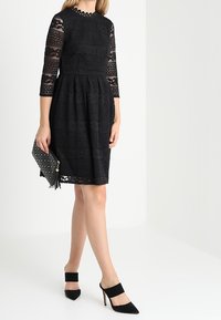 Svart spetsklänning med trekvartsärmar och rund halsringning, lagerkjol, matchad med en svart nitat väska och klackar.