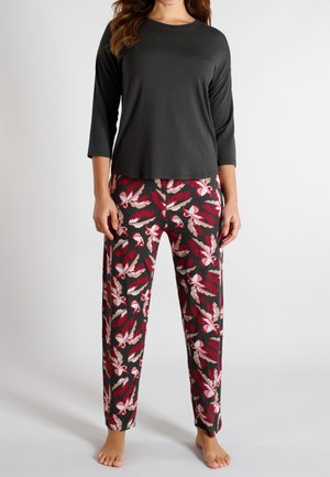 Frau trägt ein schwarzes Oberteil mit dreiviertellangen Ärmeln und lockere, gerade geschnittene Hosen mit roten, rosa und beigen Blattmustern, steht barfuß.