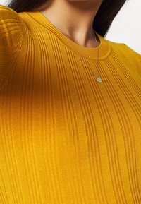 Gelbes, geripptes Strickoberteil mit Rundhalsausschnitt, subtiler Textur und enganliegendem Schnitt. Kombiniert mit einer filigranen silbernen Halskette.