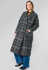 Manteau double boutonnage en mélange de laine dans un motif à carreaux gris avec de grandes poches et des revers crantés, associé à un jean en denim bleu évasé et des chaussures noires.