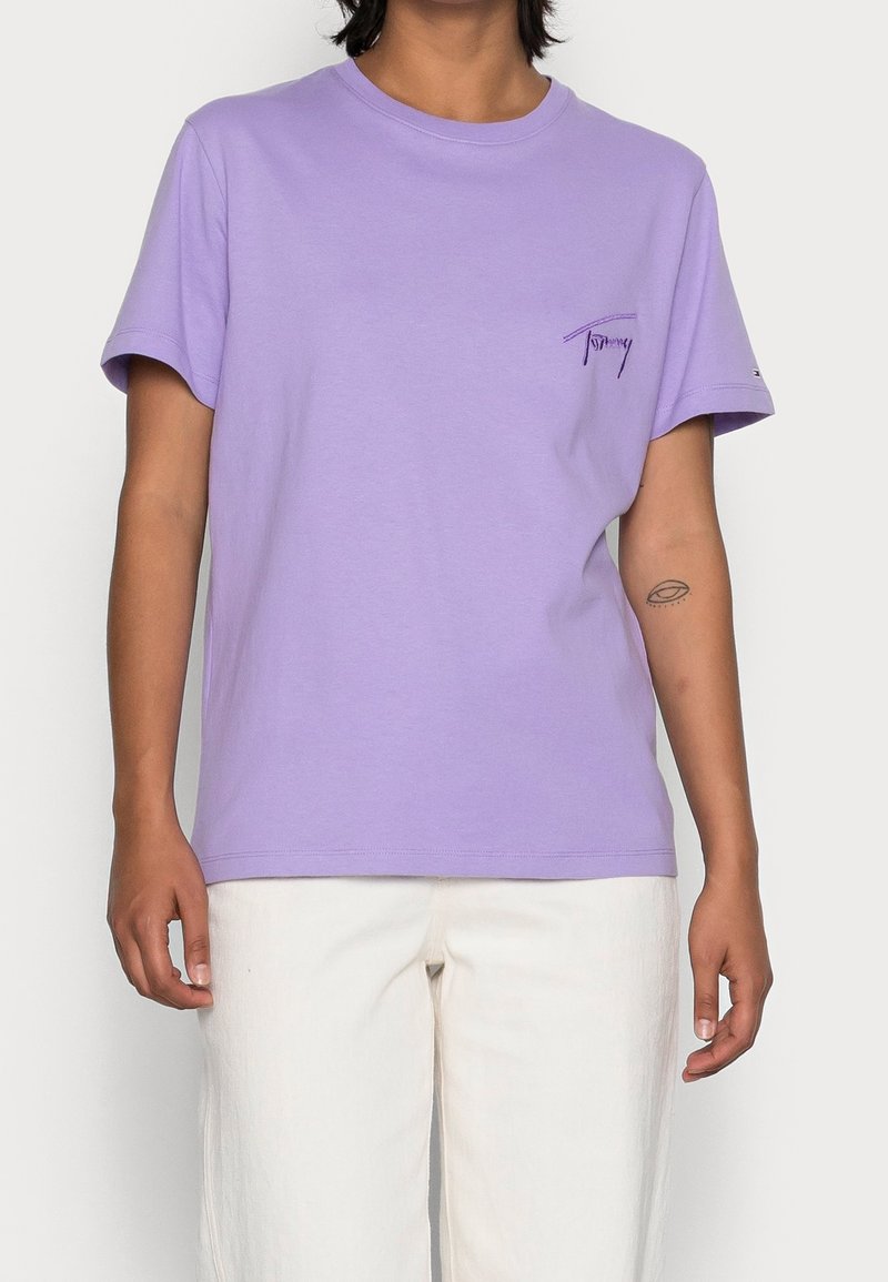 T-shirt in cotone lilla con maniche corte, scollatura tonda e un piccolo logo ricamato sul lato sinistro del petto. Abbinato a pantaloni chiari.