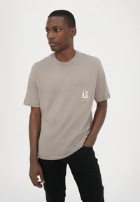T-shirt a maniche corte, marrone chiaro, con scollo rotondo e taschino sul petto con un logo bianco. Realizzato in tessuto morbido con una texture liscia.