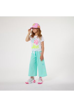 Witte t-shirt met kleurrijke grafische tekst, muntgroene wijde broek, roze pet met hartdetail en witte sneakers met roze accenten.