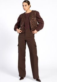 Vrouw in een bruine fuzzy trui, een sequined bruine vest, brede bruine cargobroek en bruine puntige schoenen, staande met haar handen in haar zakken.