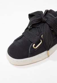 Baskets en daim noir avec un lacet noir épais et texturé, des œillets dorés et une semelle en caoutchouc blanche présentant un motif rainuré.