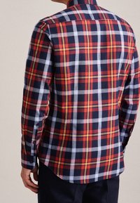 Mężczyzna w długim rękawie z koszulą w kratę w kolorach czerwonym, granatowym, białym i żółtym, pokazany od tyłu na jednolitym tle.