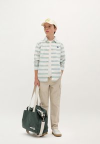 Giovano che indossa una camicia leggera a righe con bottoni, pantaloni cargo beige e sneaker bianche, mentre tiene una borsa di tela verde con un logo.