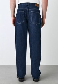 Granatowe dżinsy o luźnym kroju, z skórzaną naszkicowaną łatą na tylnej części pasa i podwójnymi szwami na kieszeniach.