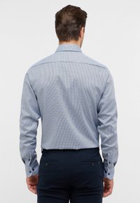 Langwijdige, blauw gemusterde shirt met een ruitpatroon. De stof is glad; het heeft een klassieke kraag en knoopmanchetten. Gecombineerd met donkere broeken.