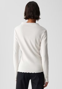 Weißes Langarmoberteil mit einem runden Ausschnitt, aus weichem Strickstoff und gewelltem Saum, kombiniert mit schwarzen Hosen.