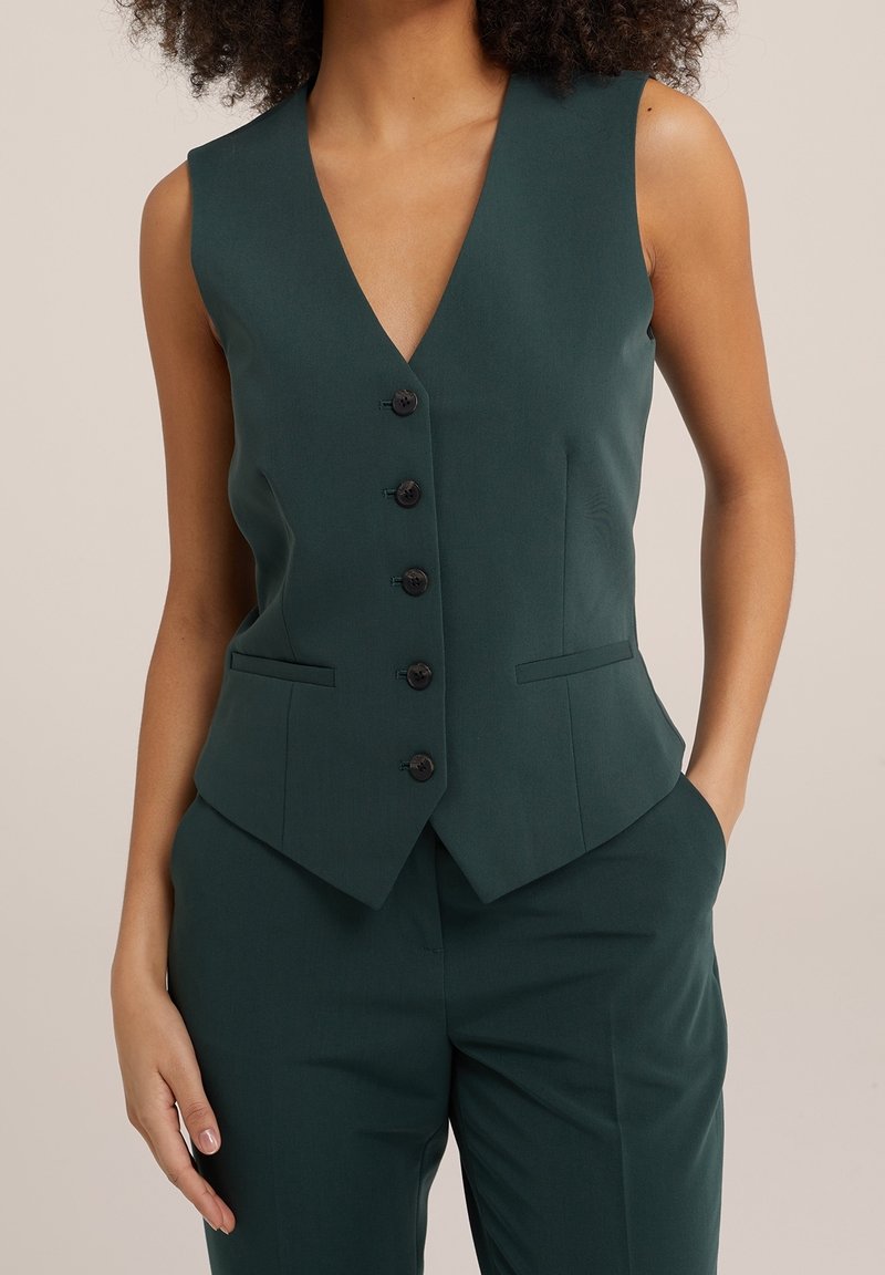 Gilet ajusté vert foncé avec un décolleté en V profond, six boutons noirs et deux poches latérales ; texture lisse et design cintré.