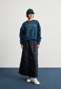 Nike kék pulóver, feltűnő fehér logóval, fekete, bokáig érő szoknyával és fehér magasszárú tornacipővel párosítva. Zöld kockás sapka kiegészítőként.