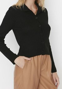 Černý pletený kardigan s hrubým pletem a límcem, s knoflíkovým zapínáním a přiléhavým střihem. Kombinovaný s kalhotami z hnědé umělé kůže.