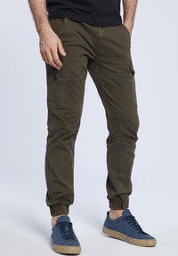 Spodnie bojówki w oliwkowym kolorze z elastycznymi mankietami, wyposażone w wiele kieszeni i gładką bawełnianą fakturę, noszone z niebieskimi trampkami.