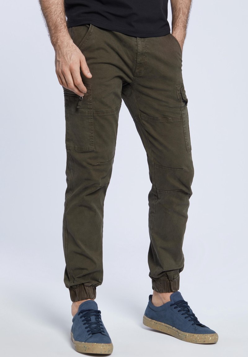 Spodnie bojówki w oliwkowym kolorze z elastycznymi mankietami, wyposażone w wiele kieszeni i gładką bawełnianą fakturę, noszone z niebieskimi trampkami.