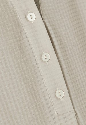Camicia beige con bottoni, caratterizzata da un tessuto testurizzato con un sottile motivo a quadretti, piccoli bottoni rotondi e una placca ordinata.