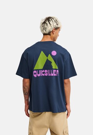 Homme aux cheveux blonds courts portant un t-shirt bleu marine avec un logo Quiksilver vert et rose dans le dos et un short cargo beige.