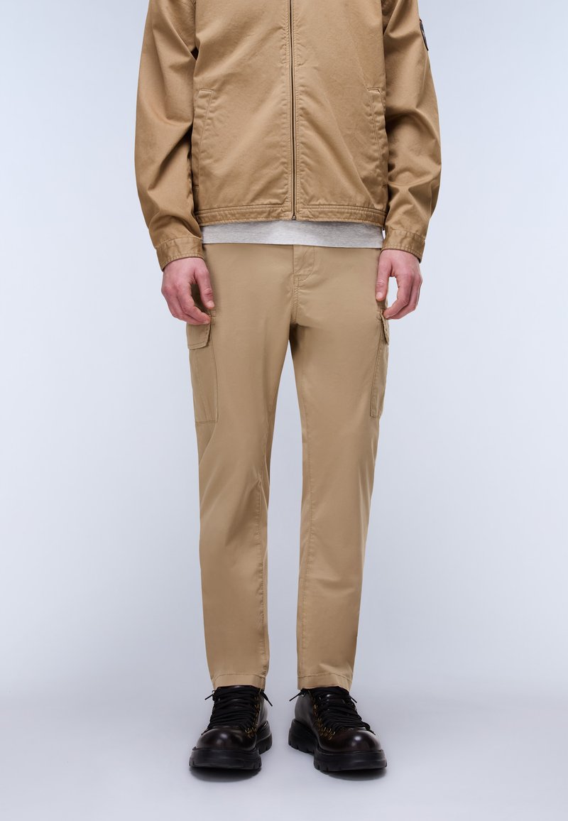 Man wearing beige cargo pants, matching beige jacket, light gray shirt, and black lace-up boots, standing against light gray background.