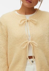 Gros plan sur une personne portant un cardigan en tricot crème fermé par deux nœuds, par-dessus un haut blanc et un pantalon blanc.