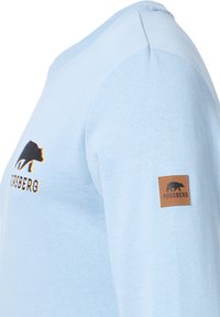 Camisa de manga larga azul claro hecha de algodón suave. Presenta un logo de oso negro en el frente y una etiqueta de cuero marrón en la manga.