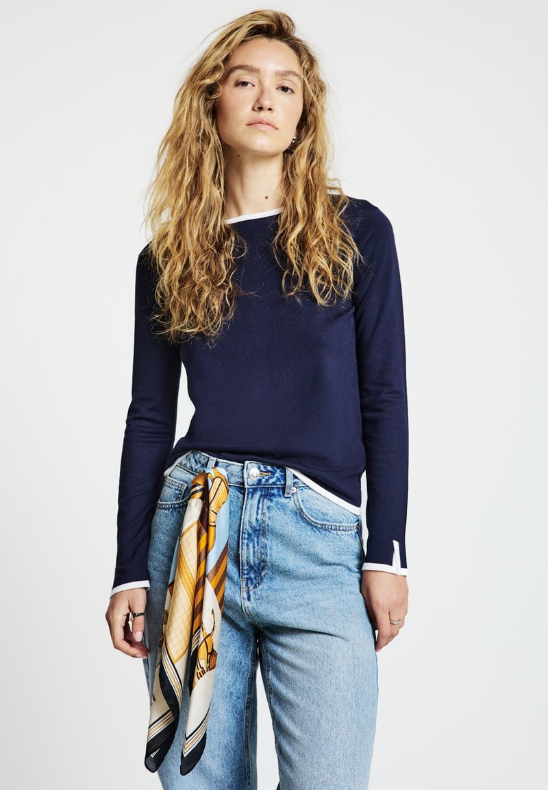 Marineblaues Langarmshirt mit weißen Akzenten am Ausschnitt und an den Bündchen, kombiniert mit hellblauen, hoch taillierten Jeans und einem gemusterten Seidenschal, der an der Taille gebunden ist.