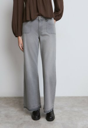 Personne portant un jean gris taille haute à jambes larges avec des ourlets effilochés, un chemisier marron à manches longues et des bottines noires, debout sur un sol en béton.