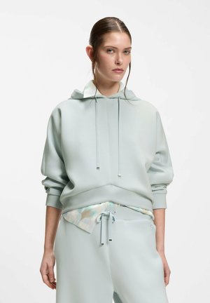 Vrouw draagt een lichtgrijze oversized hoodie en bijpassende broek met trekkoord, met een gedeeltelijk zichtbare blouse met patroon eronder.