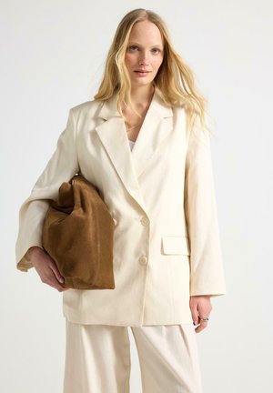 Lyshåret kvinde iført en cremefarvet dragt holder en stor brun ruskindtaske mod en ensfarvet hvid baggrund.