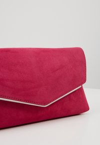 Miękka torebka kopertówka o zamszowej fakturze w czerwonym kolorze, z srebrnym obszyciem na skośnej klapie, spoczywająca na białej powierzchni na jednolitym tle.