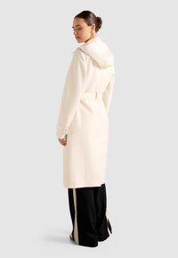 Donna che indossa un lungo trench beige con cappuccio, vita segnata da una cintura e pantaloni neri a gamba larga con strisce beige, in piedi di spalle.