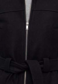 Manteau noir avec une fermeture éclair argentée partiellement ouverte montrant une couche intérieure grise, avec une ceinture en tissu nouée à la taille.