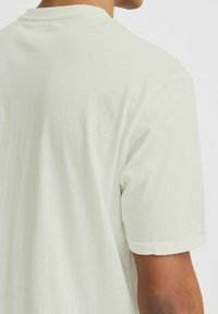 Camisa de manga corta beige claro, de tejido suave, con cuello redondo y mangas enrolladas, que presenta una textura suave con costuras mínimas.