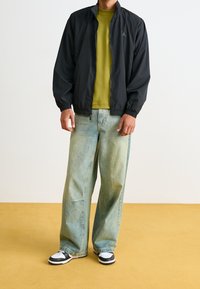 Svart bomberjacka med dragkedja och elastiska ärmslut över en grön t-shirt. Löst sittande, ljusblå jeans och svart-vita sneakers fulländar outfiten.