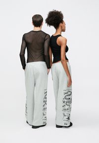 Kaks modelli seisavad seljaga vaataja poole, kandes läbipaistvast mustast kangast pika varrukaga särke ja musta tankitoppi. Mõlemal on seljas hallid dressipüksid, mille külgedel on graafilised mustrid.