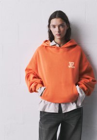 Orange oversized hoodie with a front pocket, featuring the text "Cute But Wild" in a lighter shade. Worn over a striped button-up shirt.